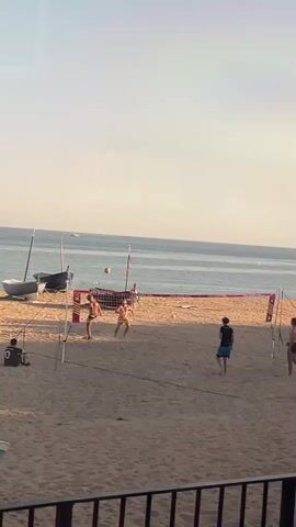 The video shows a beach volleyball game in progress. Four people are visible on the sand, two on each side of a net. They are dressed in casual beach attire. The ball is in the air, and players are actively engaged in hitting it back and forth. A person sits to the left near some parked sailboats. The ocean is in the background, and the sky is clear.