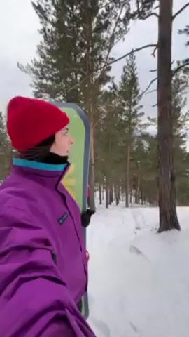 A young woman in a purple jacket and red hat walks uphill in a snowy landscape, carrying a snowboard. The terrain is covered in snow and scattered with trees. Another individual is visible further downhill, snowboarded through the trees. The sky is overcast.