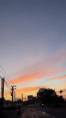 The video shows a vertical view of a road at dusk. The sky is a gradient of purple and pink clouds, with a bright orange glow near the horizon. On the left side of the road, there are utility poles with many wires. On the right side, there are trees and some palm trees. There is a billboard in the distance. A few motorcycles are visible on the road. One motorcycle is on the left side of the frame, and another is on the right side, further down the road. The road appears wet, suggesting it may have rained recently. The overall mood is peaceful and serene.