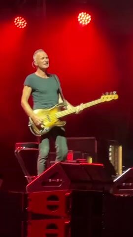 A male musician, likely Sting, performs on a stage. He is wearing a dark t-shirt and dark pants and is playing a bass guitar. He is standing on top of a stack of speakers, with red stage lights illuminating the background. The stage appears to be dark, with equipment visible in the background, including more speakers and some stage rigging. The musician is looking towards his left and appears to be singing. The video captures a live performance setting.