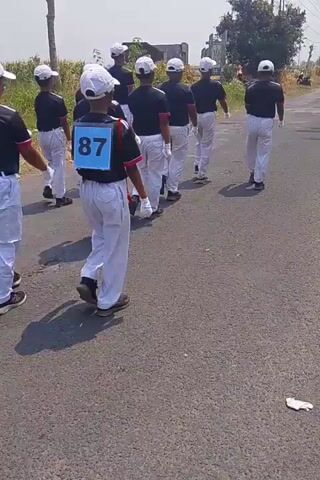 A group of boys wearing black shirts, white pants, and white hats are marching in formation on a paved road. The boy in the front right has a blue bib with the number 87 on it. In the background, there is a motorcycle and a person standing next to it. There are also trees and power lines visible. The scene appears to be outdoors on a sunny day.