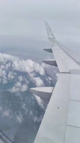 The video shows an aerial view from an airplane window. The right wing of the aircraft is prominently in the foreground, extending diagonally across the frame. Below the wing, a landscape is visible, partially obscured by fluffy white clouds. The clouds are scattered, allowing glimpses of the ground, which appears to be a mix of green terrain and possibly some signs of human habitation, such as faint lines suggesting roads or fields. The sky is overcast with a pale gray hue, indicating a cloudy day. The overall impression is one of a flight over a scenic, though not perfectly clear, landscape.