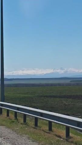 The video shows a highway on a sunny day. The road has two lanes in each direction, separated by a barrier. There are a few cars driving in the distance. In the background, there is a field and some mountains. There is a sign that reads "14" on the side of the road. There are also numerous light poles along the side of the highway.