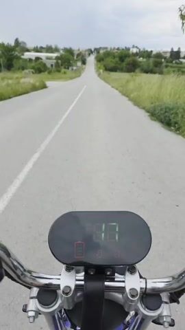 Here is a description of the video. 
The video shows a first-person perspective of someone riding an electric bike on an empty road. The road stretches straight ahead and slopes slightly uphill towards a small town. There is greenery on either side of the road, and the sky is overcast. The focus is on the bike's speedometer, which displays a gradually increasing speed in bright green digits. The battery indicator shows a nearly full charge. The chrome handlebars are visible.