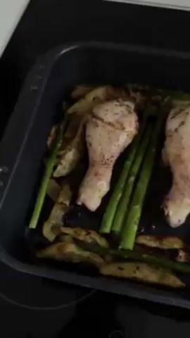 Here is a brief, detailed description of the video:

The video shows a close-up view of a black roasting pan containing chicken drumsticks, asparagus, and potato wedges. The drumsticks appear to be seasoned with herbs. The potatoes look like they have been roasted to a golden brown color with some edges appearing slightly charred. The asparagus spears are green and lie beside the chicken. The pan is placed on a black stovetop. The camera angle is slightly tilted, providing a top-down perspective of the food.