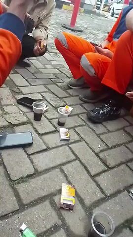 Several men, some wearing orange jumpsuits, are sitting on a brick-paved area outside. There are disposable cups of coffee, cigarettes, and phones on the ground. The men are interacting and using their phones. There is a building and a car in the background.