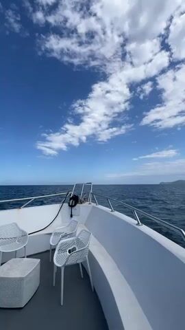 The video shows the view from the front of a white boat sailing on the open ocean. The boat has several white chairs and a small white table. The ocean is a deep blue, and in the distance, a coastline is visible. The sky is bright blue with numerous puffy white clouds. The camera slowly pans from left to right, showcasing the vastness of the ocean and the distant shore.