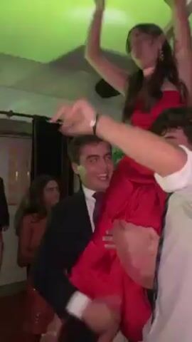 The video shows a woman in a two-piece red satin outfit being carried on the shoulders of two men. One of the men is wearing a suit jacket and the other is in a white shirt. The woman is smiling and waving her arms. There are other people visible in the background, including a woman in a sequined dress. The lighting is green. Everyone appears to be dancing and enjoying themselves.