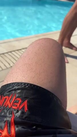 This media file captures a sunny day at a poolside resort in Calvià, Spain. The scene is dominated by a bright blue swimming pool, with people visible in and around it, enjoying the water and the warm weather. The camera focuses on close-ups of legs and swimwear, suggesting a relaxed, personal perspective of someone lounging by the pool.

In the foreground, a person's hairy leg and black swim shorts with red "VENOM" lettering are prominently displayed. To the right, another person's legs and feet are visible, also seemingly relaxing. The poolside area is paved with light-colored tiles, and a striped deck chair is partially visible, covered with a beige towel featuring a stylized eye design. A white bucket filled with ice and a green glass are on a small, round, cream-colored table, indicating drinks are being served.

As the view pans out, the context becomes clearer. The pool area extends further, with more lounge chairs and umbrellas. In the background, buildings and lush green trees suggest an urban or resort setting. The media file then shifts to a view from a balcony overlooking a coastal town. The sea is visible in the distance, with boats sailing. Buildings, some with balconies, are clustered along the coastline, with palm trees interspersed. The sky is a clear, bright blue, with a few scattered white clouds, and the sun is high, suggesting it is midday or early afternoon. A bottle of sparkling wine, likely champagne, is chilling in an ice bucket on the same table with the glasses. The overall impression is one of leisure and relaxation in a holiday destination.