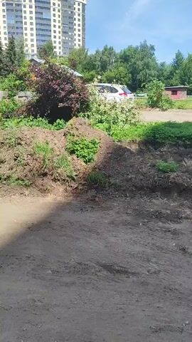 The video captures an outdoor scene on a sunny day in Shchyolkovo, Russia. The foreground is a rough, dusty ground, indicating a work-in-progress or undeveloped area. Behind this, there are piles of soil and some greenery, suggesting recent excavation or landscaping.

In the middle ground, several cars are parked. A white sedan is prominent, with a person in a light-colored shirt visible near its driver's side. Further back, another white car, possibly an SUV, is parked. A darker colored car is also present. A stack of grey concrete pavers, bound with white straps, is placed near the foreground, implying construction or renovation work. There is also a small, red-colored structure, possibly a shed or utility building, visible in the background.

The background is dominated by apartment buildings, characteristic of a residential area. One tall building is a light-colored high-rise, while another, to its right, is a multi-story building with a reddish-brown facade and numerous windows. Lush green trees are scattered throughout the scene, providing a contrast to the urban structures. The sky is blue with scattered white clouds, suggesting fair weather. The overall impression is a somewhat unkempt urban environment undergoing some form of external work, likely related to paving or landscaping. No text is visible in the video.
