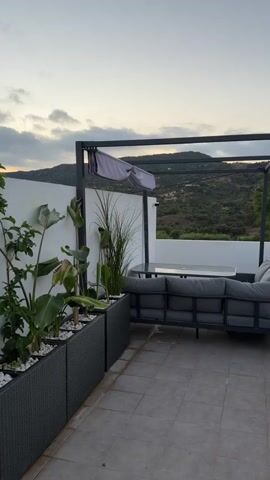 The video shows an outdoor seating area with a large gray sectional couch and a dark table underneath a black canopy. There are also planters with plants and white rocks in the foreground. In the background, there is a white wall and a view of hills and sky. The time of day appears to be dusk. There are no people or animals visible in the video.