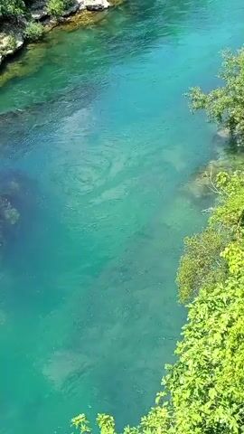 An aerial view of a clear, turquoise river flowing through a rocky, forested canyon. Lush green foliage lines the right bank, with the water exhibiting subtle currents and ripples. The sun casts a bright light on the scene, illuminating the riverbed and surrounding vegetation.