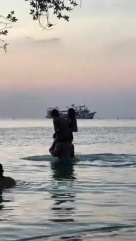 Silhouetted figures, partially submerged in the ocean, are engaged in horseplay, a child perched on an adult's shoulders. Another figure stands nearby in the water. A large boat is visible in the distance, under a sky painted with hues of sunset. A tree branch frames the top left of the shot.