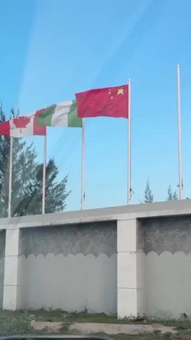 The camera films a row of flags waving on flagpoles on top of a grey wall. From left to right, the flags are the Canadian flag, the flag of Nigeria, and the flag of China. Behind the wall and the flags, there are a few small trees and a clear blue sky. The bottom of a car door is visible in the lower-left corner of the shot.
