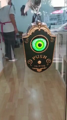 The video shows a Halloween-themed doorbell and decoration. At the beginning, a doorbell shaped like a shield with a moving green eyeball in the center hangs on a glass door. The text "PUSH TO RING" is inscribed below the eye. When a button is pushed, the eye blinks, the door makes a moaning sound and a ghastly voice says "Eeeeaaargghh." The camera shifts to reveal a severed hand hanging from the door. A person is visible in the background wearing a white shirt and dark pants. The door is in a storefront, with a view of a street outside. The interior has a light wooden floor and salon-style furniture.