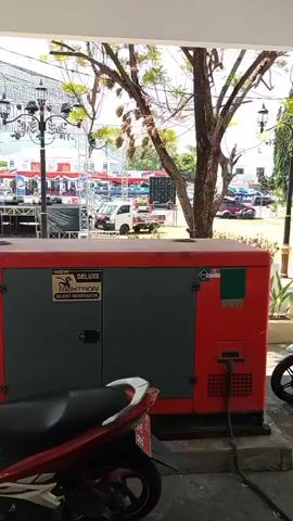 A generator is visible in the foreground, red and green in color, with a black label that reads "DELUXE SILENT GENERATOR". A motorcycle is parked to the right of the generator. In the background, a white truck is parked near a stage with sound equipment. Other vehicles and people are visible in the distance. The scene appears to be outdoors, with trees and streetlights present.