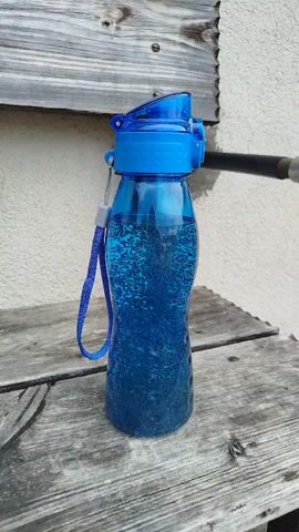 A blue water bottle filled with a blue liquid and small bubbles is placed on a weathered wooden surface. The bottle has a blue cap and a blue strap attached to its side. A light-colored wall and a wooden plank are visible in the background, along with a black object extending from the right side of the frame. The lighting suggests an outdoor setting.