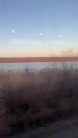 The video shows a landscape viewed from a moving vehicle.  The sky is blue with pink and orange hues near the horizon. Below the sky, a body of water extends across the frame. The water surface appears somewhat choppy.  In the foreground, there is thick vegetation, primarily consisting of dry grasses and brush. The camera moves steadily.  Dust particles and small imperfections are visible on the window through which the scene is filmed. Occasional clicks are heard.