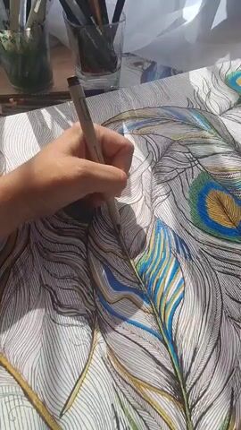 This media file shows a close-up, overhead view of a person's hand coloring a detailed drawing of peacock feathers. The drawing is on a white surface, possibly a table or a large piece of paper. The feathers are rendered with intricate black lines, and some have been partially colored with shades of blue, green, and gold. The hand, holding a fine-tipped pen or marker, is actively adding detail to one of the feathers, focusing on the outlined areas.

In the background, out of focus, are two glass containers filled with art supplies, including pencils and brushes. The lighting suggests it is daytime, with sunlight casting shadows on the drawing. The setting appears to be a well-lit indoor space, potentially an art studio or a home where someone is engaged in a creative activity. The overall mood conveyed is one of concentration and artistic engagement. There is no visible text in the media file. The location context of Kempelevo, Russia, provides a geographical setting for this scene.