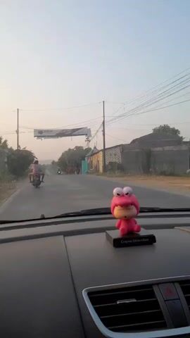 This is a first-person perspective video taken from inside a car. A pink, frog-shaped bobblehead is visible on the dashboard. The car is on a road, and the view through the windshield shows a rural or semi-rural landscape. There are trees lining the road, and some buildings on the right side. Several people on motorcycles are traveling in the same direction as the car. A banner is strung across the road overhead, displaying text in what appears to be Indonesian. The sky is a pale, hazy blue, suggesting it might be early morning or late afternoon.