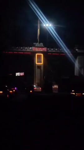 A nighttime shot focuses on an illuminated archway with red lettering reading "रायगड" (Raygad) above a framed portrait of a man. The archway is topped with decorative battlements and a flagpole with a flag. Bright lights cast beams across the scene. Below the arch, traffic lights and vehicles are visible, including a white car and motorcycles. The overall impression is of an entrance to a town or landmark at night.