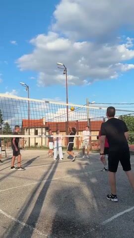 An outdoor volleyball game is in progress on a paved court under a partly cloudy blue sky. A yellow and blue volleyball is airborne above the net, which spans the midground of the scene. Approximately eight individuals, appearing to be young adults or teenagers, are distributed on both sides of the net. On the left side, a player in a white shirt is in the process of hitting or setting the ball. Several other players in various colored shirts and shorts are positioned around the court. On the right side, a player in a black shirt and shorts stands with their back to the camera, observing the play, while another player in a red shirt watches the ball. Shadows from the players and the net are cast on the court. Two streetlights on tall poles and residential buildings are visible in the background.