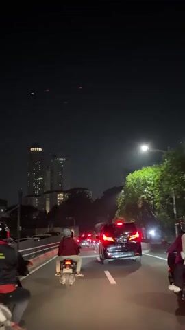 The video shows a nighttime scene of traffic on a road. In the foreground, a person on a scooter is riding in the right lane, facing away from the camera. The scooter's taillight is illuminated red. To the right of the scooter, a black SUV with its red taillights illuminated is traveling in the same direction. Several other vehicles are visible further down the road, with their headlights and taillights creating streaks of light in the darkness. Tall buildings with illuminated windows are visible in the background. Trees line the right side of the road, and a streetlamp provides light. The sky is dark, and there appear to be some red lights or reflections in the upper part of the frame. The road has white lane markings.