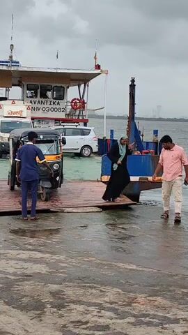 The video shows a ferry boat with the name AVANTIKA and the registration number NIV 030082 on its side. An auto-rickshaw is positioned on the ramp of the ferry. A person in a dark blue uniform stands to the left of the auto-rickshaw, facing it. Another person, a woman wearing a hijab and a black dress, is walking up the ramp from the right side of the frame. A man in a pink shirt and light-colored pants is standing on the ramp to the right of the woman, facing her and gesturing. The ferry ramp is wooden and appears wet. In the background, other vehicles, including a white SUV and a white van, are visible on the ferry. The sky is cloudy. The scene appears to be taking place at a ferry terminal.