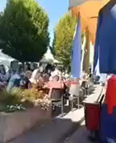 This is a video of an outdoor market or fair. There are several people in the scene, some of whom are sitting at tables. There are also tents and stalls set up, and clothing items are hanging on display. Two trees with full green foliage are visible in the background, and the sky is clear and blue, indicating a sunny day. There is a building with a blue awning on the left side of the frame.