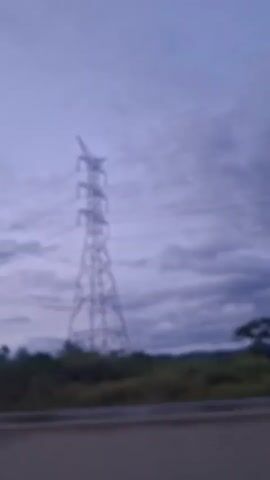 A transmission tower stands tall against a cloudy sky. The tower is made of steel lattice and supports power lines that stretch across the frame. The ground below the tower is covered in vegetation and trees. The scene appears to be captured from a moving vehicle, as indicated by the slight blur and the perspective of the road in the foreground. The lighting suggests it is either early morning or late evening.