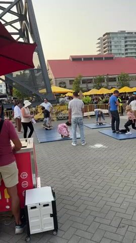 The video shows an outdoor plaza with a brick-paved surface. Several people are engaged in activities on mats laid out on the ground. In the foreground, a person in a maroon shirt and khaki shorts is partially visible, standing near a red and white cart. To the left, a large, dark metal structure, possibly part of a bridge or a building, dominates the upper left of the frame. A red umbrella is also visible.

In the middle ground, a group of people are spread across several blue mats. On one mat, a woman in a beige top and black pants is standing next to a young child in a pink dress, who is on a balance board. To their right, a man in a striped shirt and jeans stands facing away from the camera. Further to the right, a man in a blue shirt and jeans is on another mat, and a young girl in a pink shirt and white pants is also on a mat, bent over. Other individuals are scattered on mats or standing nearby.

In the background, there are yellow umbrellas and tables, suggesting an outdoor dining or market area. A building with a red roof is visible, and a tall modern building is seen in the far background. The sky is overcast, and the lighting suggests it is late afternoon or early evening. The overall atmosphere appears to be a casual outdoor event or activity.