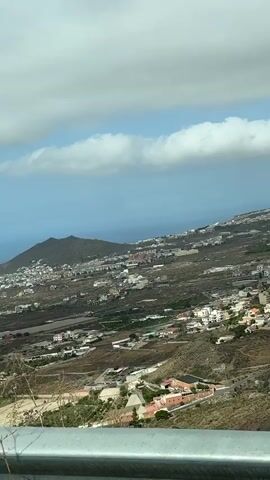 A drone flies over a scenic landscape in Tenerife, Spain, capturing a panoramic view of the island. The drone starts by showcasing a tall, flowering agave plant, its flower stalk reaching towards the sky. The camera then pans to reveal a sprawling town nestled on a hillside, with houses of various colors dotting the landscape. In the distance, a large, dormant volcano dominates the horizon, its slopes covered in vegetation. The drone continues to move, offering glimpses of the coastline and the vast Atlantic Ocean. Signs indicating "Sentinela" and a campsite are visible, suggesting recreational opportunities in the area. The footage is characterized by its bright, natural lighting and clear skies, with fluffy white clouds adding to the picturesque scenery. The drone's smooth movements and high-resolution capture create an immersive experience for the viewer, highlighting the natural beauty and charm of Tenerife.