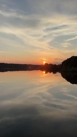 This is a vertical, wide shot of a serene lake at sunset. The sky is a warm gradient of orange, pink, and pale blue with wispy clouds. The sun, a bright orange orb, is partially obscured by silhouetted trees on the horizon. The sun's reflection creates a shimmering orange and yellow path on the still water. The opposite shoreline is also silhouetted with trees. The overall mood is peaceful and tranquil.