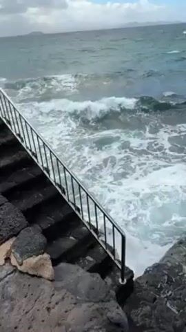 The video shows waves crashing against a rocky shoreline. A set of stone steps with a metal railing leads down to the water. The sea is choppy and a light blue color. In the distance, a white building stands on the shoreline and beyond it is a beach. The sky is cloudy. There are no people visible in the video.