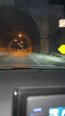 The video shows a point-of-view shot from inside a vehicle traveling through a tunnel. The tunnel is illuminated by yellow lights on the ceiling at regular intervals. The walls of the tunnel appear to be constructed of corrugated metal or concrete. The road is paved and has yellow lane markers. There are other vehicles visible in the distance ahead. The dashboard of the vehicle is partially visible in the foreground. The scene is dark, with the exception of the tunnel lighting and the headlights of the vehicle.