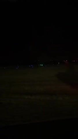 A dimly lit airport tarmac is visible at night. Various runway lights, some green and some blue, are scattered across the field. A red light is visible in the distance, and a larger red light is seen in the mid-ground on the right side of the frame. The sky is completely dark. The camera appears to be looking out from a window.