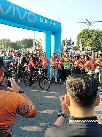 An outdoor cycling event is depicted from behind two male spectators, who are both recording with their phones. A large blue inflatable archway, labeled "VIVO" and "ELITE CELL," spans a paved road. Numerous cyclists on various bicycles, including mountain bikes, are actively moving forward under and past the archway. The cyclists are dressed in casual attire, with some wearing helmets. On the sides of the road and near the archway, a crowd of people observes the event. Several individuals wearing orange and brown patterned shirts are visible near the archway, appearing to interact with or cheer on the cyclists. The scene is brightly lit by daylight.