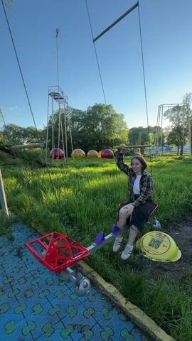 A young woman, likely in her late teens or early twenties, is shown in a park, riding a unique swing-like amusement device. She is fair-skinned with short brown hair, wearing a plaid shirt, black pants, and off-white sneakers. She is sitting on a red seat of the apparatus, holding onto a bar overhead, and smiling. The device has a red, geometrically-shaped wheel/ crank mechanism near the base. The ground is a mix of blue interlocking tiles and green grass, with other playground equipment visible in the background, including a tower structure and large, colorful, spherical objects. The video is shot outdoors during daylight under a clear blue sky, with trees visible in the background.