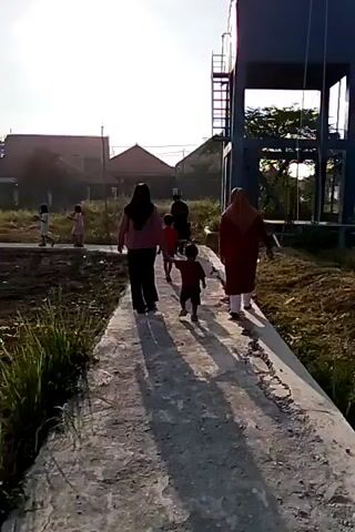 A group of people, including adults and children, are walking along a concrete path. The path appears to be cracked and uneven. In the background, there is a large, blue water tower, some houses, and overgrown vegetation. The sun is shining brightly, casting long shadows. The overall impression is of a rural or semi-rural setting.