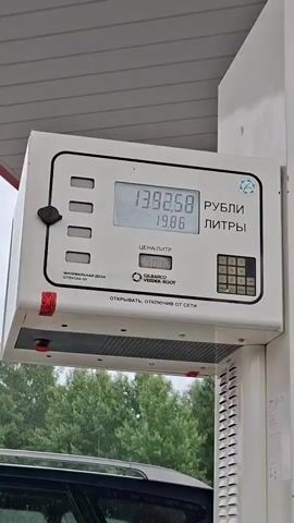 The video shows a gas pump display with changing numbers. The top number increases from 1409.82 to 1542.64, representing rubles. The second number increases from 20.18 to 22.00, representing liters. The words "РУБЛИ" (rubles) and "ЛИТРЫ" (liters) are visible. A person is partially visible at the bottom of the frame, interacting with the pump or vehicle. The environment appears to be outdoors, with trees in the background and a white structure of the gas station canopy overhead.