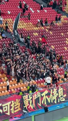 The video shows a large group of soccer fans, most of whom are wearing black jerseys, cheering in the stands of a stadium. They are waving flags, pumping their fists in the air, and appear to be chanting. The stadium seating is a mix of red and orange, and there are people seated and standing throughout the section. A large banner in front of the group features Chinese characters in gold. Security personnel are also visible near the front of the section.