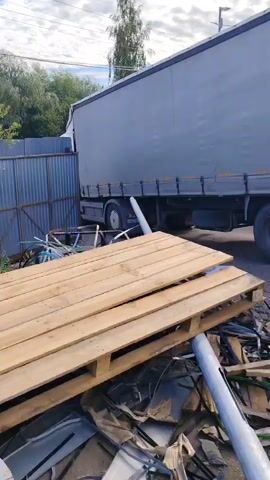 The video shows a large grey semi-trailer truck with its back doors open. The truck is parked on a dirt or gravel surface, and in the foreground, there is a pile of wooden pallets and various discarded materials, including what appears to be scrap metal and plastic. To the left of the truck is a corrugated metal fence. Behind the truck, trees and buildings are visible, and the sky is partly cloudy, suggesting it is daytime. The overall scene appears to be an outdoor storage or industrial area, possibly a scrapyard or a loading dock. There are no people visible in the video.

On the back of the truck, a license plate is visible, with Cyrillic lettering and numbers, including "BB 205 ZP". The open doors of the trailer reveal the interior of the cargo space. The lighting suggests it is daytime, and the weather seems fair with some clouds. The location is identified as Dolgoprudny, Russia. The overall impression is one of a utilitarian, possibly industrial setting where goods are transported or stored.