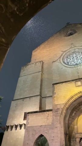 The video displays a static shot of the illuminated facade of a large, historic stone church at night. The church is constructed from light-colored stone blocks, giving it a weathered appearance.

A prominent, tall, and deep arched entrance dominates the lower central part of the facade. The archway is made of darker stone, and recessed within it are large, dark wooden doors, possibly with glass panels, upon which two large posters are affixed. The left poster features an image of a person and the partially visible text "Francisci." The right poster has a more abstract design with illegible text.

Above the main entrance, there is a rectangular stone plaque or crest. Further up, a large, ornate circular rose window with intricate tracery is visible, brightly lit and standing out against the dark stone. At the very top of the gable, a small, simple cross is silhouetted against the night sky. On either side of the main facade, there are sturdy stone buttresses. To the left of the main entrance, a smaller, darker arched opening is also visible at ground level.

The entire facade is bathed in warm, artificial yellow light, highlighting the architectural details and contrasting with the dark night sky. In the extreme top left of the frame, a dark, curved stone archway is partially visible, suggesting the camera is positioned beneath another structure. The ground in front of the church appears to be paved with dark material. The sky above is very dark, and there's a reflective, textured surface at the very top of the frame, possibly indicating a wet surface or rain. Faint ambient sounds are present in the background.