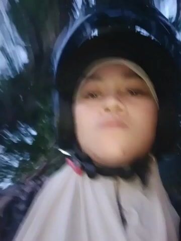A close-up, blurry video shows a person wearing a helmet and a headscarf while riding on a motorcycle. The person's face is visible, and they appear to be looking directly at the camera. The background is out of focus and shows trees and lights, suggesting the video was taken at night or in low light conditions. The person is wearing a light-colored garment that covers their neck and shoulders. The video is shaky, indicating movement, likely from the motorcycle.