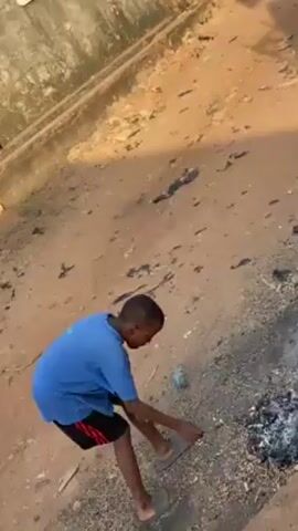 A young boy is standing in a dirt area with a partially burnt pile of debris or material next to him. He is wearing a blue t-shirt, black shorts with red stripes on the side, and slides. In his right hand he is holding what appears to be a lit match or lighter, and he is looking at it. The background includes a concrete ledge and building or wall. There is loose trash on the ground near the boy. The area appears to be a backyard or similar open space.