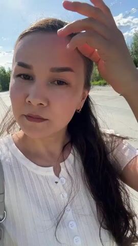 The video shows a young woman standing on a concrete sidewalk beside a parking lot. She has long dark hair, light skin, and is wearing a white button-down shirt and a light-colored skirt that's visible at her midriff. She has a light-colored shoulder bag with a chain strap.  In the background, there are cars parked in a lot. Trees are visible further behind. The sky is bright and sunny. The woman adjusts her hair and looks directly at the camera. The video seems to be a selfie.