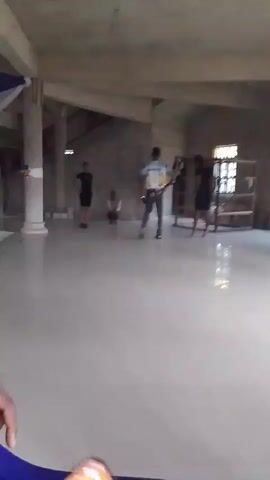 The video shows a large, unfinished room with a tiled floor. There are four people in the room. A person dressed in black shorts and a black shirt stands near a pillar on the left side of the frame. Another person sits on the floor between the first person and a man standing near the center of the room, facing away from the camera. This man is wearing jeans and a colorful short-sleeved shirt. To his right stands a woman in a black dress and heels. In the background, there's a staircase and a wooden shelf. Someone holds a phone in the foreground, recording the scene. The overall setting appears to be an informal gathering or rehearsal.