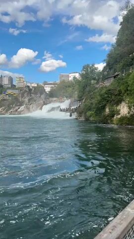 The video captures a wide view of a powerful waterfall cascading down a rocky cliff face under a bright blue sky with scattered white clouds. The water in the foreground is a deep green, rippling with white foam, suggesting a strong current. On the right side of the frame, a lush green, tree-covered hillside rises steeply from the water. Several people are visible standing on a viewing platform or pathway built into the side of the cliff near the waterfall, observing the spectacle. In the background, across the river and beyond the waterfall, modern buildings, including multi-story structures and some with yellow accents, are situated on a higher elevation, nestled among trees. The overall scene depicts a natural wonder integrated with a developed urban environment.