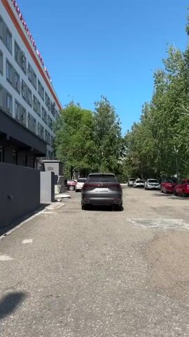 A vertical, full-shot video captures a side view of a large, multi-story building with a striped facade of white, orange, and pink. A dark gray car is parked on a paved road in front of the building. The road leads past a row of green trees and continues into the distance. There are other cars parked along the road. The sky is a clear blue. In the foreground, a person with dark hair is partially visible. The camera moves slightly, showing more of the building and the surrounding trees. Another person, wearing a light green outfit and carrying a bag, walks away from the camera. A dark blue car is parked nearby, and a white car is behind it. The camera then pans to the right, showing more of the building and trees, and finally the video ends.