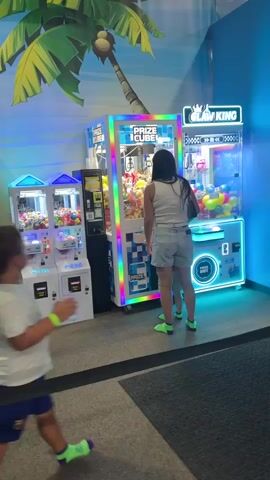 A young boy in a white t-shirt and blue shorts walks past a row of arcade prize claw machines. He is wearing bright green shoes. To his right, a woman in a white tank top and denim shorts stands with her back to the camera, watching a large claw machine filled with colorful stuffed animals. She is also wearing bright green shoes. A smaller child, wearing an orange shirt and gray shorts, stands next to her, facing the claw machine. The claw machines are brightly lit with colorful LEDs. The background features a mural of a palm tree and a tropical scene.